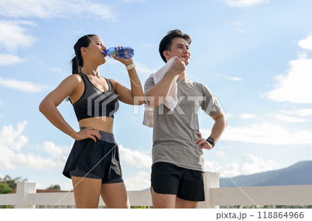 A tired, sweaty couple in sportswear is resting together after a long run in a park on a sunny day. 118864966