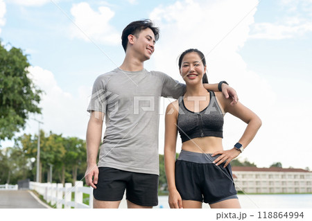 A lovely, active Asian couple in sportswear stands in a park, smiling at the camera. 118864994