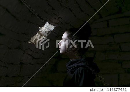 Young adult in a basement looking in a small window. A beam of light falls on her face. Portrait of the young girl in the dungeon with dark background. Young adult in a basement looking in a small window. A beam of light falls on her face. Portrait of the young girl in the dungeon with dark background. 118877481