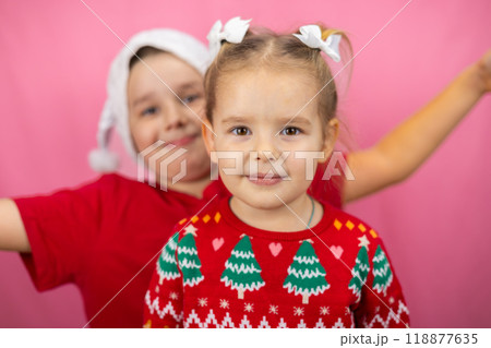 Cute baby girl posing for a portrait in a Christmas sweater while her brother peeks out from behind making a funny face. Two cute kids in Christmas clothes on pink isolated background. Cute baby girl posing for a portrait in a Christmas sweater while her brother peeks out from behind making a funny face. Two cute kids in Christmas clothes on pink isolated background. 118877635
