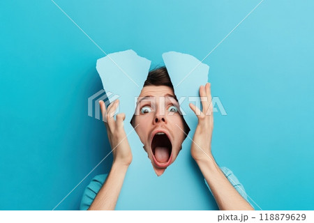A young man expressing shock and surprise through a torn blue wall effect in a studio setting A young man expressing shock and surprise through a torn blue wall effect in a studio setting 118879629