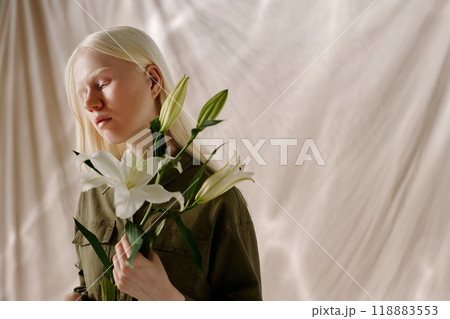 Young woman with long white hair holding fresh white lily flowers posing for camera in studio decorated with beige drapery Young woman with long white hair holding fresh white lily flowers posing for camera in studio decorated with beige drapery 118883553