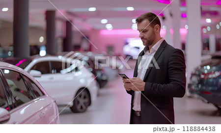 Salesman in elegant suit using smartphone at car showroom. Professional salesperson working in modern car dealership. Modern technology. 118884438
