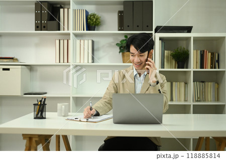 Handsome businessman talking on mobile phone and using laptop at office desk Handsome businessman talking on mobile phone and using laptop at office desk 118885814