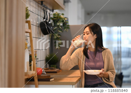 Young asian woman holding a plate of food and drinking milk while sitting at kitchen counter 118885840