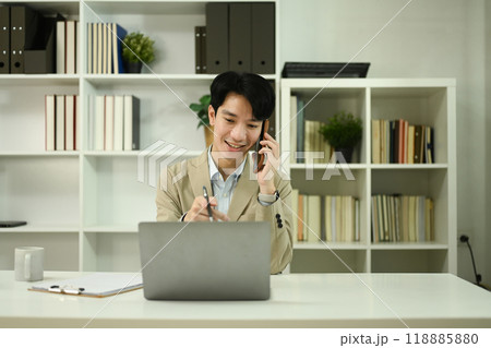 Smiling male financial counselor talking on a mobile phone and using laptop in office Smiling male financial counselor talking on a mobile phone and using laptop in office 118885880