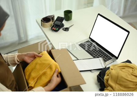 Young woman packing a cardboard box in front of laptop with blank screen. E-commerce and Online selling concept 118886026