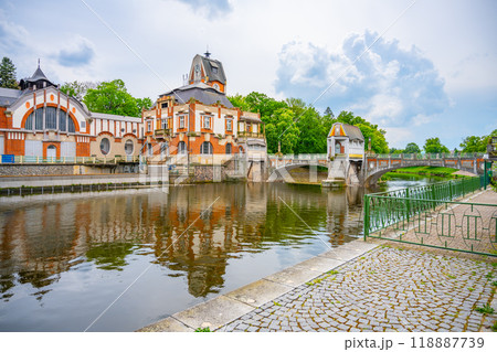 The historic Art Nouveau Hucak hydroelectric power station stands beside a tranquil river in Hradec Kralove, Czechia, showcasing intricate design and surrounded by lush greenery under a cloudy sky. 118887739