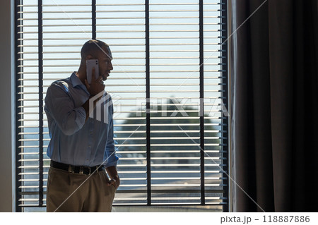 Man wearing shirt posing near big window in office and talking on mobile phone 118887886