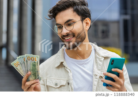 Happy Indian man counting money dollar cash, use smartphone calculator app in urban city street 118888340