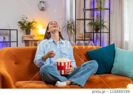 Excited young woman sitting on couch eating popcorn and watching interesting TV serial film game Excited young woman sitting on couch eating popcorn and watching interesting TV serial film game 118888399