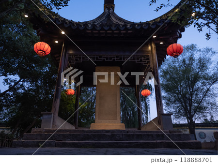 A traditional Chinese pavilion illuminated by lanterns during twilight in a serene garden setting with lush trees 118888701