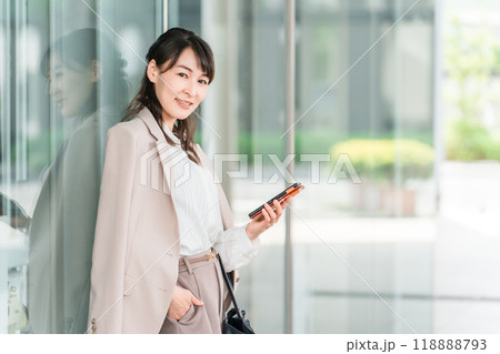 オフィスビル・商業施設の前でスマホを見ながら待ち合わせするビジネスウーマン・ワーキングマザー オフィスビル・商業施設の前でスマホを見ながら待ち合わせするビジネスウーマン・ワーキングマザー 118888793