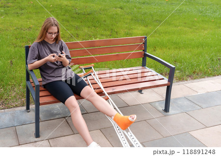 Teenage patient with a leg cast resting on a park bench, leveraging her smartphone for social interaction and video calls during her rehabilitation journey in the peaceful summer setting 118891248
