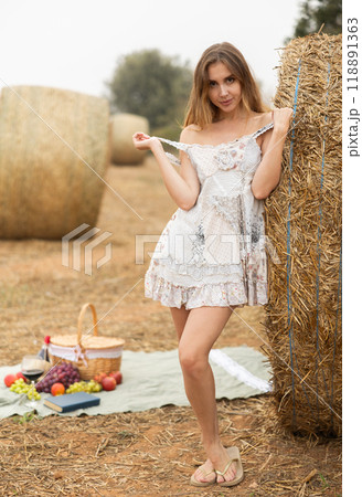 Sexy woman in white dress having picnic standing by hay bales in farm field Sexy woman in white dress having picnic standing by hay bales in farm field 118891363