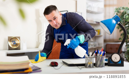 Cleaning man in overalls wipes dust from table in office Cleaning man in overalls wipes dust from table in office 118891855