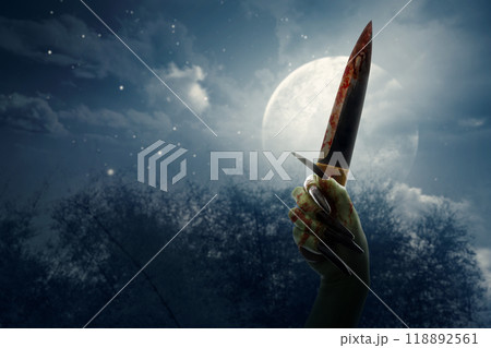 Witch hand with long black nail holding knife in a dark forest with full moon in the background. The knife is bloody and hand is green. Evil, satan, zombie or monster hand 118892561