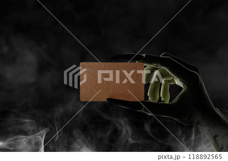 Witch hand with long black nail holding piece of brown paper with smoke on dark background. Blank piece of brown paper for copy space. Evil, satan, zombie or monster hand Witch hand with long black nail holding piece of brown paper with smoke on dark background. Blank piece of brown paper for copy space. Evil, satan, zombie or monster hand 118892565