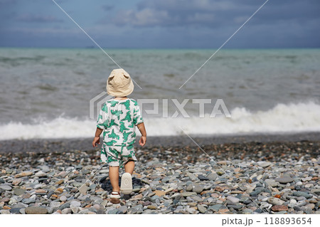 A toddler in a cute outfit and a sunhat walks along a rocky beach near the ocean as waves crash nearby. The scene captures the child's adventurous spirit and the natural beauty of the stormy A toddler in a cute outfit and a sunhat walks along a rocky beach near the ocean as waves crash nearby. The scene captures the child's adventurous spirit and the natural beauty of the stormy 118893654