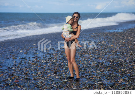 A young mother in sunglasses holds her toddler while standing on a pebble beach by the ocean. The child wears a sunhat and swimsuit, and the waves crash behind them, capturing a joyful family moment. A young mother in sunglasses holds her toddler while standing on a pebble beach by the ocean. The child wears a sunhat and swimsuit, and the waves crash behind them, capturing a joyful family moment. 118893888