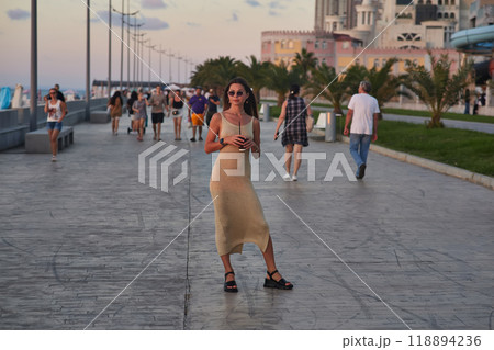 A stylish woman in a light summer dress walks confidently along a busy promenade at sunset, holding a cup of coffee. The background features palm trees, people, and seaside buildings, creating a relax A stylish woman in a light summer dress walks confidently along a busy promenade at sunset, holding a cup of coffee. The background features palm trees, people, and seaside buildings, creating a relax 118894236