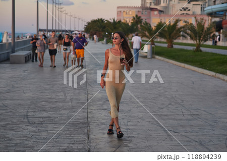 A stylish woman in a light summer dress walks confidently along a busy promenade at sunset, holding a cup of coffee. The background features palm trees, people, and seaside buildings, creating a relax 118894239