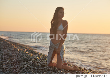 A young woman in a bikini kneels on a rocky beach at sunset. The soft evening light highlights her hair and silhouette as gentle waves wash up on the shore. Peaceful moment by the sea. A young woman in a bikini kneels on a rocky beach at sunset. The soft evening light highlights her hair and silhouette as gentle waves wash up on the shore. Peaceful moment by the sea. 118895312
