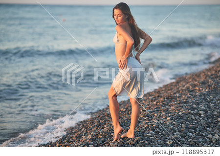 A young woman in a bikini kneels on a rocky beach at sunset. The soft evening light highlights her hair and silhouette as gentle waves wash up on the shore. Peaceful moment by the sea. A young woman in a bikini kneels on a rocky beach at sunset. The soft evening light highlights her hair and silhouette as gentle waves wash up on the shore. Peaceful moment by the sea. 118895317