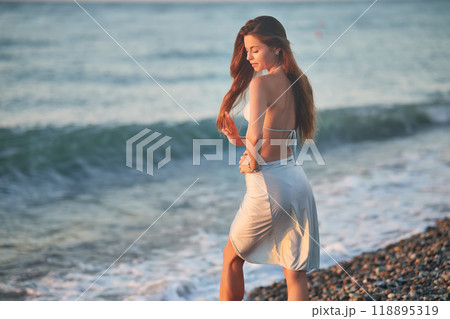 A young woman in a bikini kneels on a rocky beach at sunset. The soft evening light highlights her hair and silhouette as gentle waves wash up on the shore. Peaceful moment by the sea. 118895319