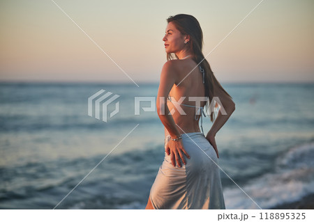 A young woman in a bikini kneels on a rocky beach at sunset. The soft evening light highlights her hair and silhouette as gentle waves wash up on the shore. Peaceful moment by the sea. A young woman in a bikini kneels on a rocky beach at sunset. The soft evening light highlights her hair and silhouette as gentle waves wash up on the shore. Peaceful moment by the sea. 118895325
