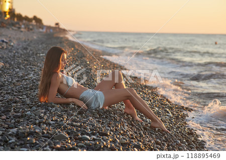 A woman relaxes on a rocky beach at sunset, wearing a bikini and cover-up skirt. The warm golden light of the setting sun reflects on the ocean, creating a serene, peaceful, and scenic atmosphere. 118895469