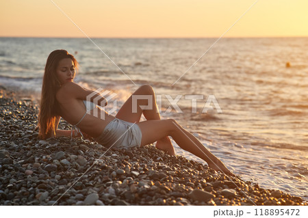 A woman relaxes on a rocky beach at sunset, wearing a bikini and cover-up skirt. The warm golden light of the setting sun reflects on the ocean, creating a serene, peaceful, and scenic atmosphere. 118895472