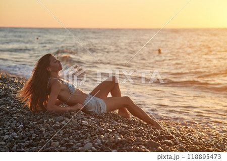 A woman relaxes on a rocky beach at sunset, wearing a bikini and cover-up skirt. The warm golden light of the setting sun reflects on the ocean, creating a serene, peaceful, and scenic atmosphere. A woman relaxes on a rocky beach at sunset, wearing a bikini and cover-up skirt. The warm golden light of the setting sun reflects on the ocean, creating a serene, peaceful, and scenic atmosphere. 118895473
