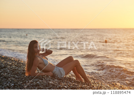 A woman relaxes on a rocky beach at sunset, wearing a bikini and cover-up skirt. The warm golden light of the setting sun reflects on the ocean, creating a serene, peaceful, and scenic atmosphere. A woman relaxes on a rocky beach at sunset, wearing a bikini and cover-up skirt. The warm golden light of the setting sun reflects on the ocean, creating a serene, peaceful, and scenic atmosphere. 118895474