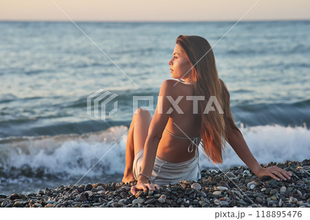 A woman relaxes on a rocky beach at sunset, wearing a bikini and cover-up skirt. The warm golden light of the setting sun reflects on the ocean, creating a serene, peaceful, and scenic atmosphere. A woman relaxes on a rocky beach at sunset, wearing a bikini and cover-up skirt. The warm golden light of the setting sun reflects on the ocean, creating a serene, peaceful, and scenic atmosphere. 118895476
