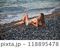 A woman relaxes on a rocky beach at sunset, wearing a bikini and cover-up skirt. The warm golden light of the setting sun reflects on the ocean, creating a serene, peaceful, and scenic atmosphere. 118895478