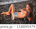 A woman relaxes on a rocky beach at sunset, wearing a bikini and cover-up skirt. The warm golden light of the setting sun reflects on the ocean, creating a serene, peaceful, and scenic atmosphere. 118895479