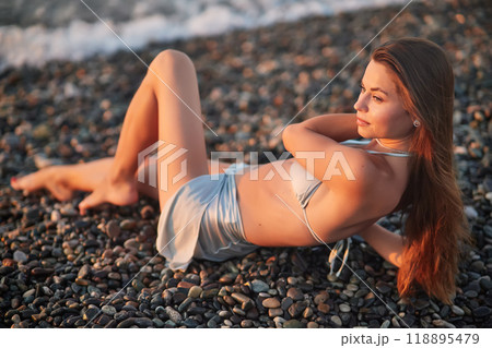 A woman relaxes on a rocky beach at sunset, wearing a bikini and cover-up skirt. The warm golden light of the setting sun reflects on the ocean, creating a serene, peaceful, and scenic atmosphere. 118895479