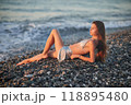 A woman relaxes on a rocky beach at sunset, wearing a bikini and cover-up skirt. The warm golden light of the setting sun reflects on the ocean, creating a serene, peaceful, and scenic atmosphere. 118895480