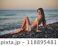 A woman relaxes on a rocky beach at sunset, wearing a bikini and cover-up skirt. The warm golden light of the setting sun reflects on the ocean, creating a serene, peaceful, and scenic atmosphere. 118895481