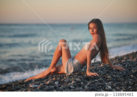 A woman relaxes on a rocky beach at sunset, wearing a bikini and cover-up skirt. The warm golden light of the setting sun reflects on the ocean, creating a serene, peaceful, and scenic atmosphere. 118895481