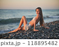A woman relaxes on a rocky beach at sunset, wearing a bikini and cover-up skirt. The warm golden light of the setting sun reflects on the ocean, creating a serene, peaceful, and scenic atmosphere. 118895482