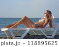A young woman relaxes on a beach lounge chair by the ocean, sipping a refreshing drink under the warm sun. The serene coastal scenery and clear blue water capture the essence of a perfect summer day. 118895625