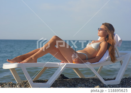 A young woman relaxes on a beach lounge chair by the ocean, sipping a refreshing drink under the warm sun. The serene coastal scenery and clear blue water capture the essence of a perfect summer day. 118895625