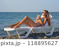 A young woman relaxes on a beach lounge chair by the ocean, sipping a refreshing drink under the warm sun. The serene coastal scenery and clear blue water capture the essence of a perfect summer day. 118895627