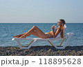 A young woman relaxes on a beach lounge chair by the ocean, sipping a refreshing drink under the warm sun. The serene coastal scenery and clear blue water capture the essence of a perfect summer day. 118895628