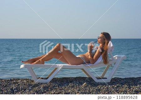 A young woman relaxes on a beach lounge chair by the ocean, sipping a refreshing drink under the warm sun. The serene coastal scenery and clear blue water capture the essence of a perfect summer day. A young woman relaxes on a beach lounge chair by the ocean, sipping a refreshing drink under the warm sun. The serene coastal scenery and clear blue water capture the essence of a perfect summer day. 118895628