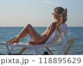 A young woman relaxes on a beach lounge chair by the ocean, sipping a refreshing drink under the warm sun. The serene coastal scenery and clear blue water capture the essence of a perfect summer day. 118895629