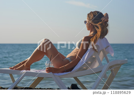 A young woman relaxes on a beach lounge chair by the ocean, sipping a refreshing drink under the warm sun. The serene coastal scenery and clear blue water capture the essence of a perfect summer day. 118895629