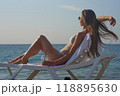 A young woman relaxes on a beach lounge chair by the ocean, sipping a refreshing drink under the warm sun. The serene coastal scenery and clear blue water capture the essence of a perfect summer day. 118895630